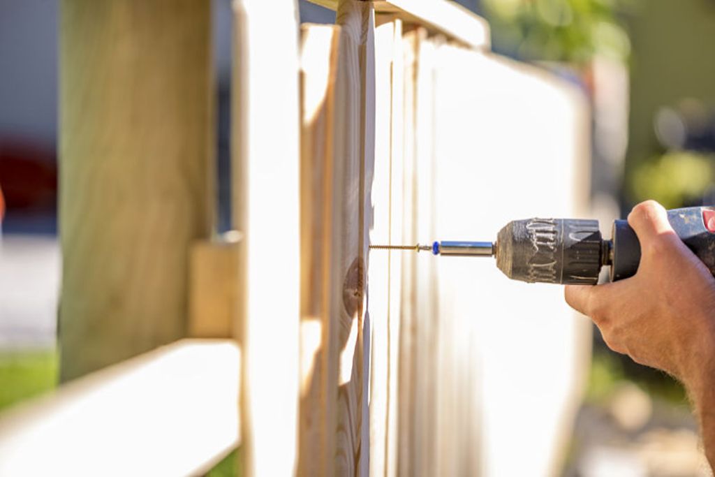 Man erecting a wooden fence outdoors using a handheld electric drill to drill a hole to attach an upright plank, close up of his hand and the tool in a DIY concept.